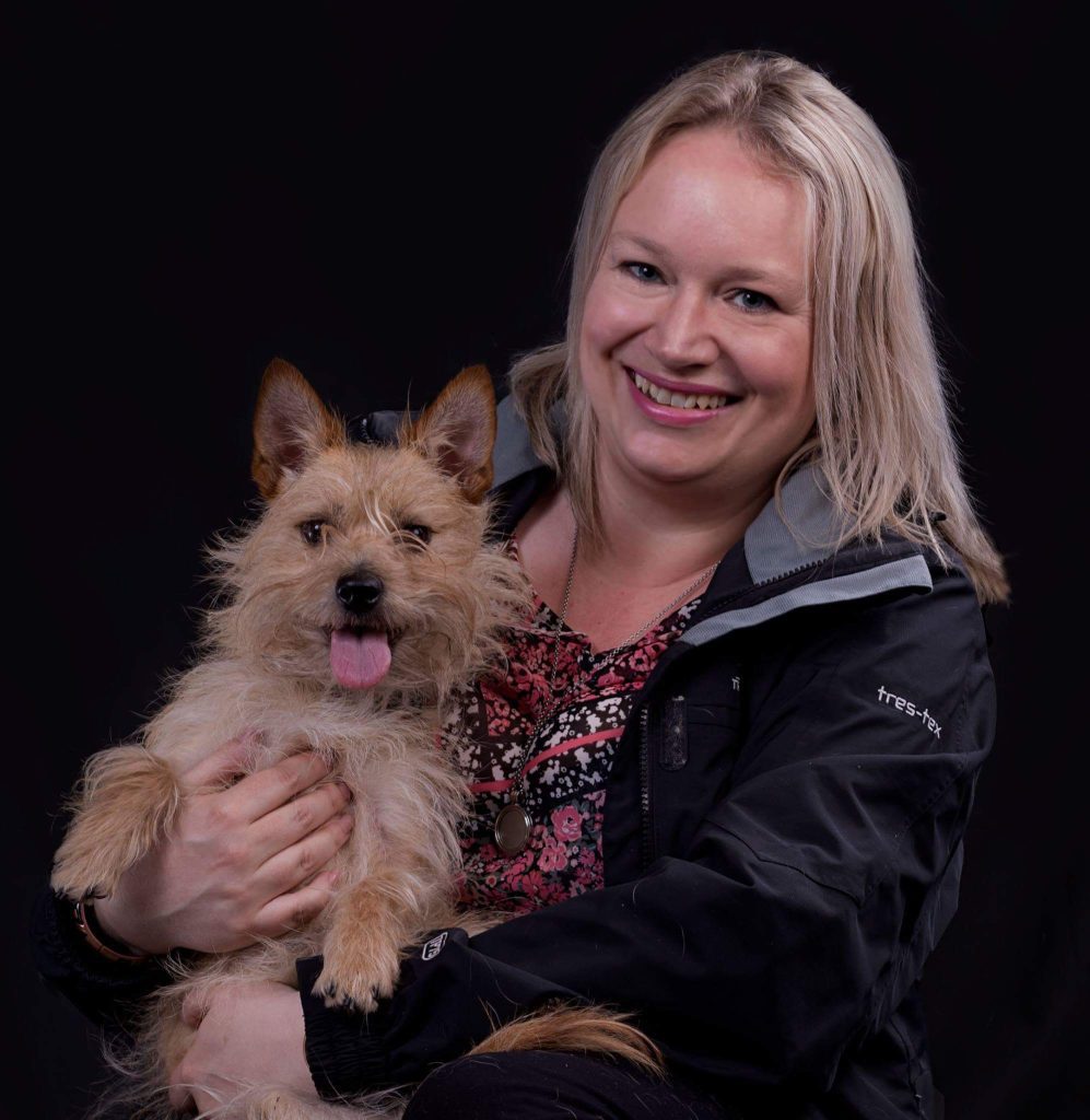 caroline mitchell sits holding her small terrier dog on her knee who has her tongue out as she looks at the camera