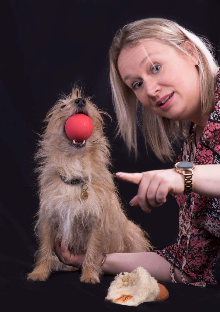 Caroline Mitchell and her small brown terrier. Caroline is pointing to the camera and the dog is sitting beside her holding a large red ball in her mouth 